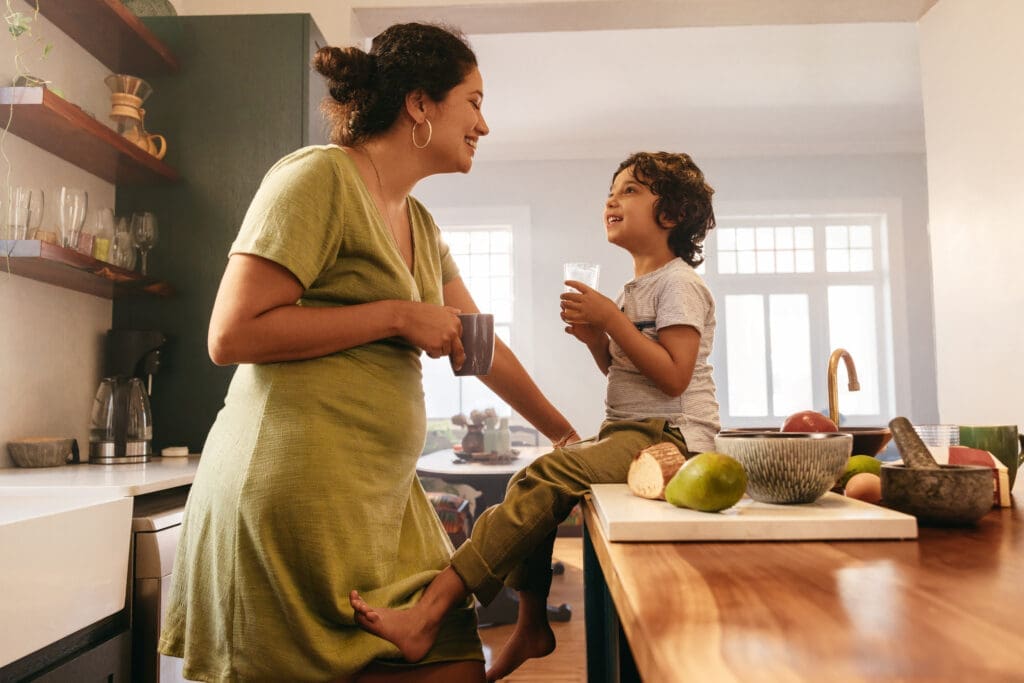 Happy mother and son smiling at each other in the kitchen Happy mother and child enjoying a calm morning in a bright, modern kitchen. The mother leans on the counter with a coffee mug while her son sits nearby with a glass of water, surrounded by fresh fruit and natural light — representing family comfort, cleanliness, and pest-free living.