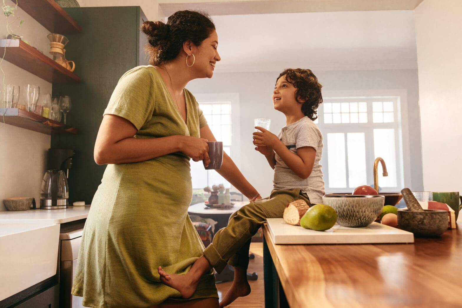 Happy mother and child enjoying a calm morning in a bright, modern kitchen. The mother leans on the counter with a coffee mug while her son sits nearby with a glass of water, surrounded by fresh fruit and natural light — representing family comfort, cleanliness, and pest-free living.